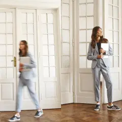 Dwie kobiety w eleganckich, **casual**owych garniturach. Jedna idzie, druga stoi, obie trzymają laptopy.