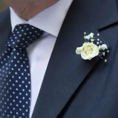 Elegancka butonierka co to? Delikatna biała róża z gipsówką zdobi ciemną marynarkę i granatowy krawat w kropki.
