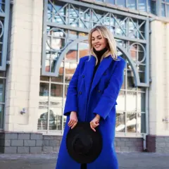 Uśmiechnięta blondynka w płaszczu w kolorze kobaltowym, trzymająca czarny kapelusz, na tle nowoczesnej architektury.