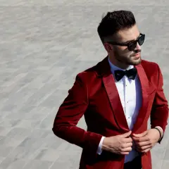 Elegancki mężczyzna w czerwonej, aksamitnej bonżurce i okularach przeciwsłonecznych poprawia marynarkę.