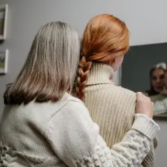 Dwie kobiety, jedna z rudymi włosami splecionymi w warkocz, druga z siwymi, patrzą w lustro. Zastanawiają się, czy toner pokryje siwe włosy.
