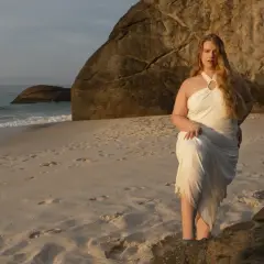 Kobieta o krągłych kształtach w białej sukni na plaży, z morzem i skałami w tle.