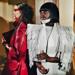 Dwie modelki w awangardowych kreacjach haute couture. Jedna w czerwonej, łuskowej marynarce, druga w białej z frędzlami.