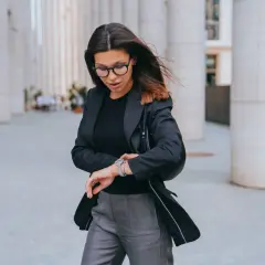 Elegancka kobieta w czarnej marynarce i szarych spodniach sprawdza zegarek. Idealny zestaw, by wiedzieć, jaka marynarka do szarych spodni pasuje najlepiej.