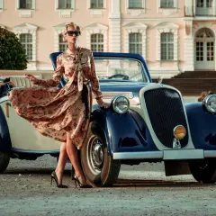 Elegancka kobieta w kwiecistej sukience i szpilkach przy zabytkowym kabriolecie. Stylowe lata 20. w pełnej krasie.