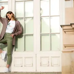 Kobieta w brązowym kardiganie i zielonych spodniach pozuje przed drzwiami.