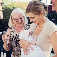 Elegancka mama karmi dziecko na chrzcinach. Babcia z kieliszkiem wina i dziadek patrzą z uśmiechem.