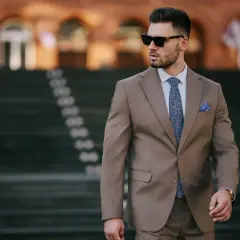 Elegancki mężczyzna w brązowym garniturze i okularach przeciwsłonecznych, z wzorzystym krawatem i poszetką, idzie po schodach.