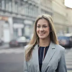 Uśmiechnięta kobieta w marynarce na tle miasta. Jej pozytywne renee opinie o życiu są widoczne w promiennej twarzy.