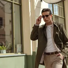 Mężczyzna w stylowym płaszczu w kolorze khaki, który to kolor jest idealny na wiosenne dni, poprawia okulary przeciwsłoneczne.