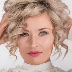 Kobieta z kręconymi blond włosami, która nonszalancko układa je dłońmi. Jej spojrzenie jest hipnotyzujące, a usta pomalowane na różowo.