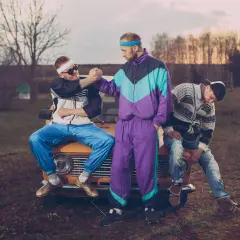 Trzech mężczyzn w dresach i swetrach, uosabiających modę lat 90., pozuje na tle starego samochodu.