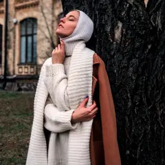 Kobieta w białej balaklawie i swetrze opiera się o drzewo.