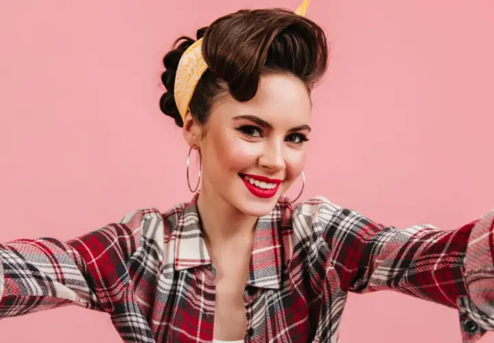 Uśmiechnięta kobieta w stylu pin-up, z burzą brązowych włosów i żółtą bandaną, w koszuli w kratę, robi selfie.