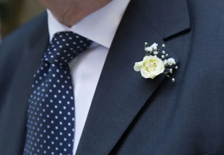 Elegancka butonierka co to? Delikatna biała róża z gipsówką zdobi ciemną marynarkę i granatowy krawat w kropki.