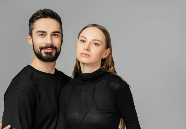 Para, która wygląda jakby mogła nosić ubrania unisex. Mężczyzna z brodą i kobieta z długimi włosami, oboje w czarnych, prostych ubraniach.