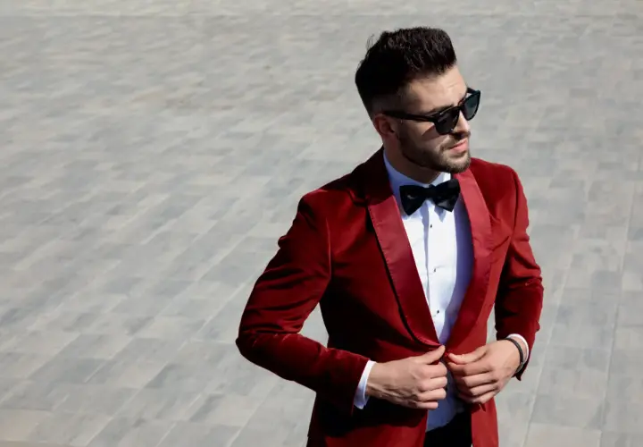 Elegancki mężczyzna w czerwonej, aksamitnej bonżurce i okularach przeciwsłonecznych poprawia marynarkę.