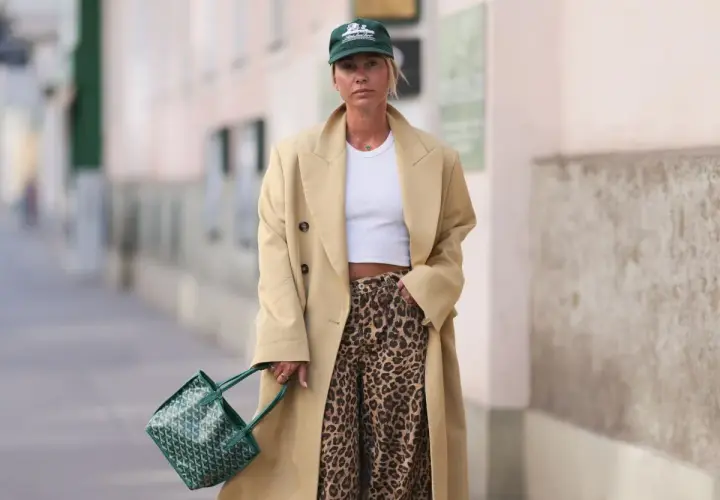Modna kobieta w beżowym płaszczu, białym topie i spodniach w panterkę. Zielona torebka i czapka dopełniają stylizację.