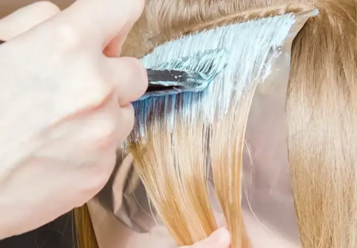 Dłoń z pędzlem nakłada jasnoniebieski rozjaśniacz na pasma blond włosów. Czas ekspozycji jest kluczowy, by uzyskać pożądany efekt.
