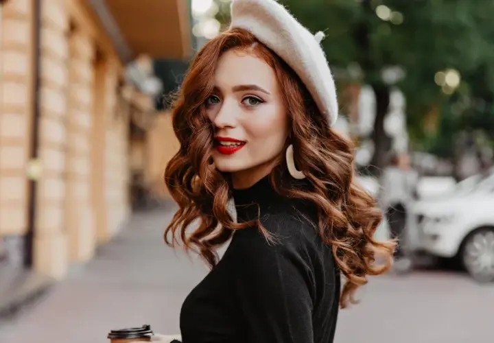 Rudowłosa kobieta w beretce i czarnym golfie, z burzą falowanych włosów i czerwoną szminką, trzyma kubek kawy.