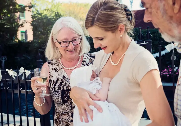 Elegancka mama karmi dziecko na chrzcinach. Babcia z kieliszkiem wina i dziadek patrzą z uśmiechem.