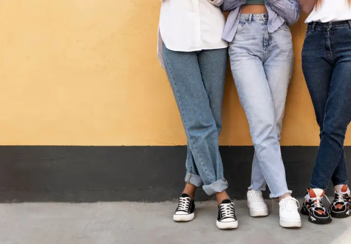 Trzy osoby w jeansach, w tym jeansy straight, stoją przy żółtej ścianie.