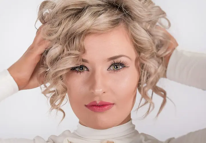 Kobieta z kręconymi blond włosami, która nonszalancko układa je dłońmi. Jej spojrzenie jest hipnotyzujące, a usta pomalowane na różowo.