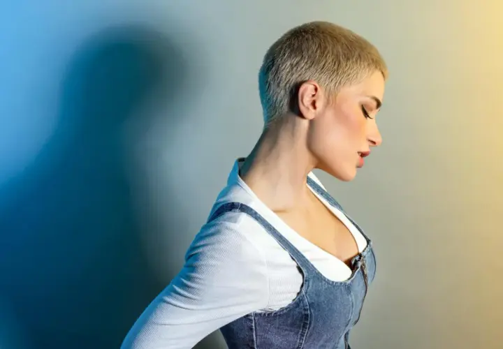 Kobieta z odważną fryzurą buzz cut, w jeansowej sukience na ramiączkach i białej bluzce.