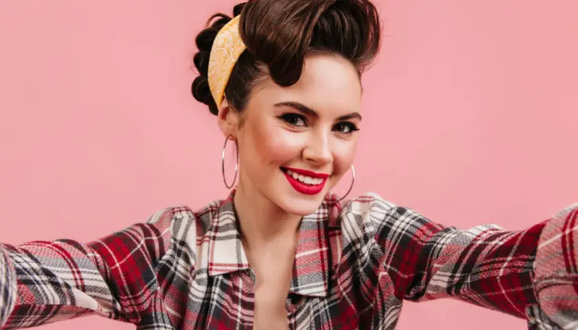 Uśmiechnięta kobieta w stylu pin-up, z burzą brązowych włosów i żółtą bandaną, w koszuli w kratę, robi selfie.