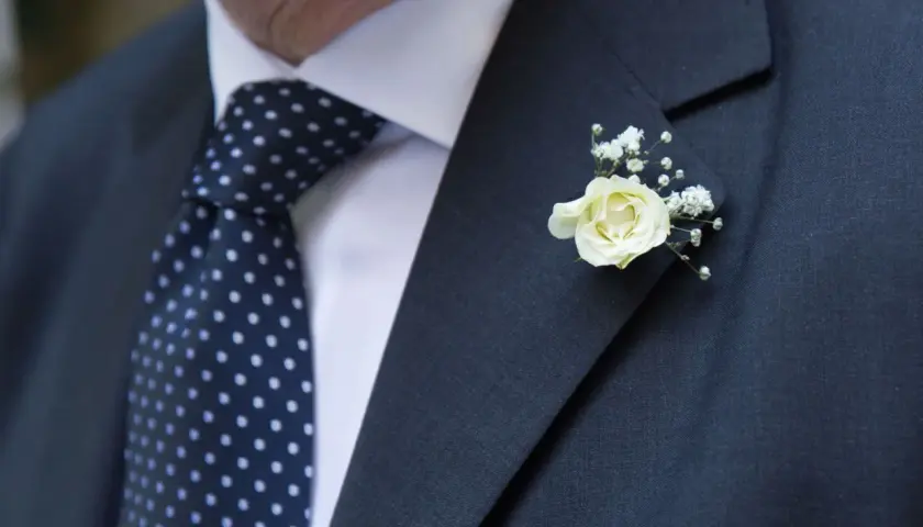 Elegancka butonierka co to? Delikatna biała róża z gipsówką zdobi ciemną marynarkę i granatowy krawat w kropki.