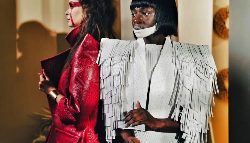 Dwie modelki w awangardowych kreacjach haute couture. Jedna w czerwonej, łuskowej marynarce, druga w białej z frędzlami.