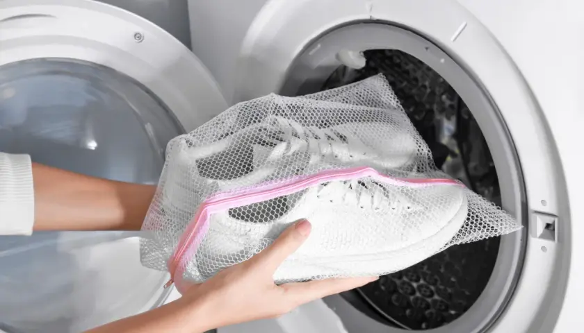 Ręce wkładają białe buty sportowe w siatkowej torbie do pralki. Pranie butów w pralce to szybki sposób na odświeżenie ulubionych sneakersów.