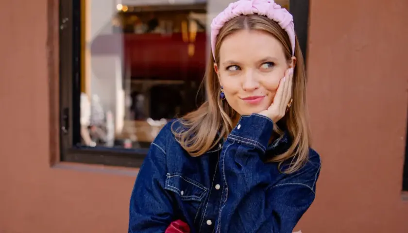 Dziewczyna w jeansowej kurtce i różowej opasce na głowie, uśmiecha się lekko, emanując stylem clean girl aesthetic.