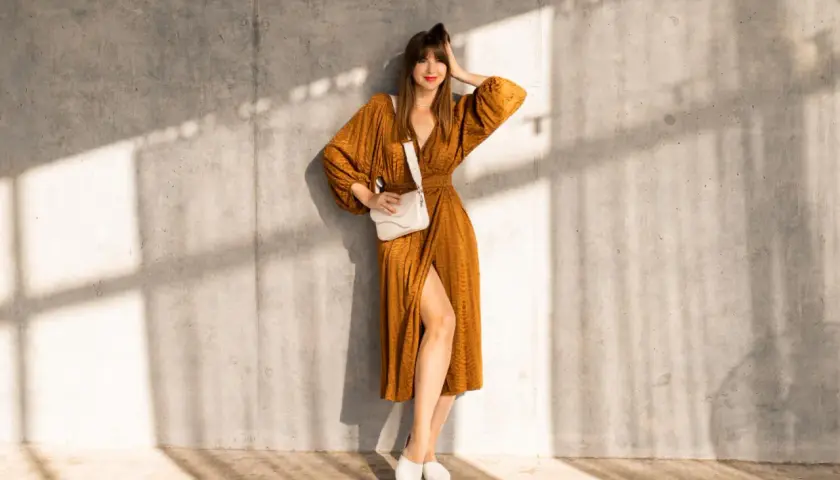 Elegancka kobieta w brązowej sukience i białych butach, oparta o betonową ścianę, z białą torebką na ramieniu.