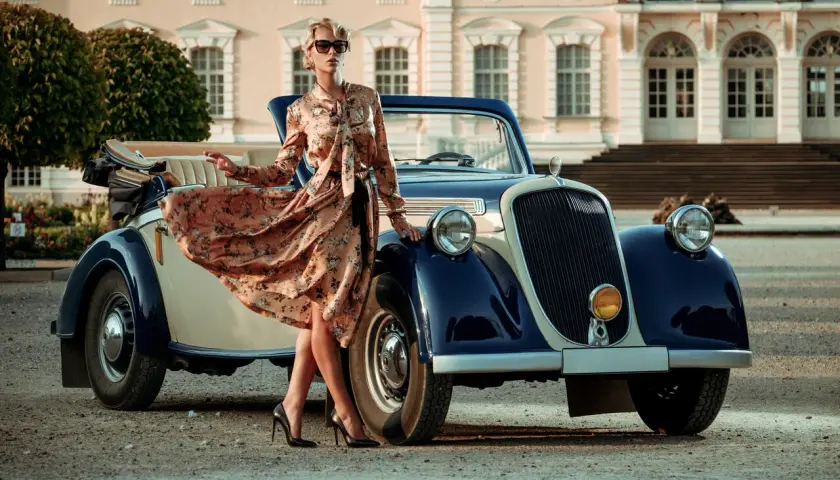 Elegancka kobieta w kwiecistej sukience i szpilkach przy zabytkowym kabriolecie. Stylowe lata 20. w pełnej krasie.