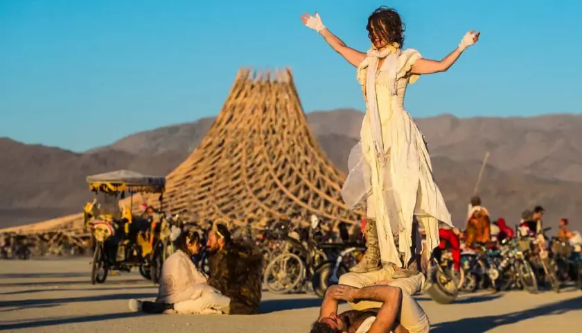 Akrobaci na pustyni podczas Burning Man. W tle charakterystyczna konstrukcja i tłum ludzi.