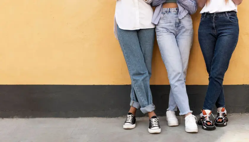 Trzy osoby w jeansach, w tym jeansy straight, stoją przy żółtej ścianie.
