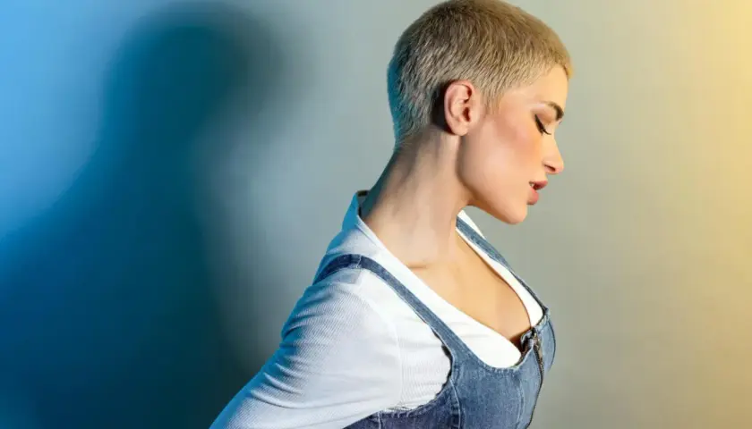 Kobieta z odważną fryzurą buzz cut, w jeansowej sukience na ramiączkach i białej bluzce.