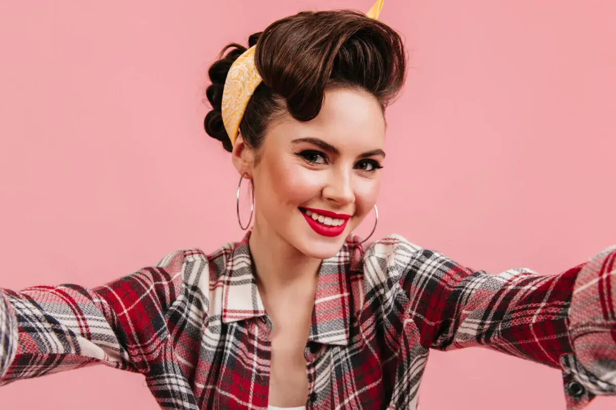 Uśmiechnięta kobieta w stylu pin-up, z burzą brązowych włosów i żółtą bandaną, w koszuli w kratę, robi selfie.