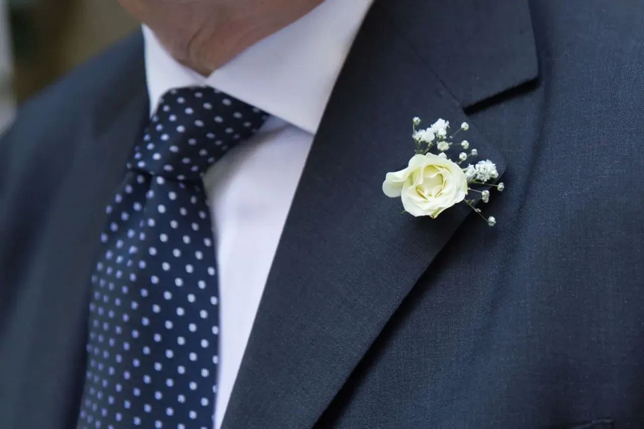 Elegancka butonierka co to? Delikatna biała róża z gipsówką zdobi ciemną marynarkę i granatowy krawat w kropki.