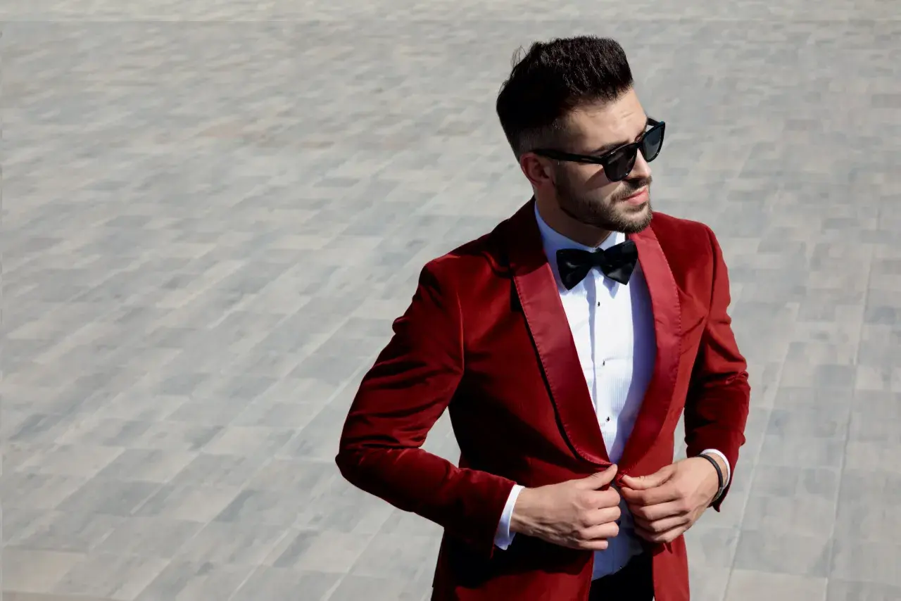 Elegancki mężczyzna w czerwonej, aksamitnej bonżurce i okularach przeciwsłonecznych poprawia marynarkę.