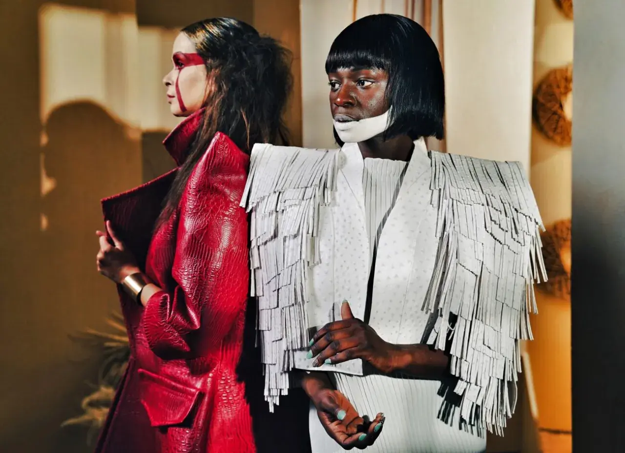 Dwie modelki w awangardowych kreacjach haute couture. Jedna w czerwonej, łuskowej marynarce, druga w białej z frędzlami.