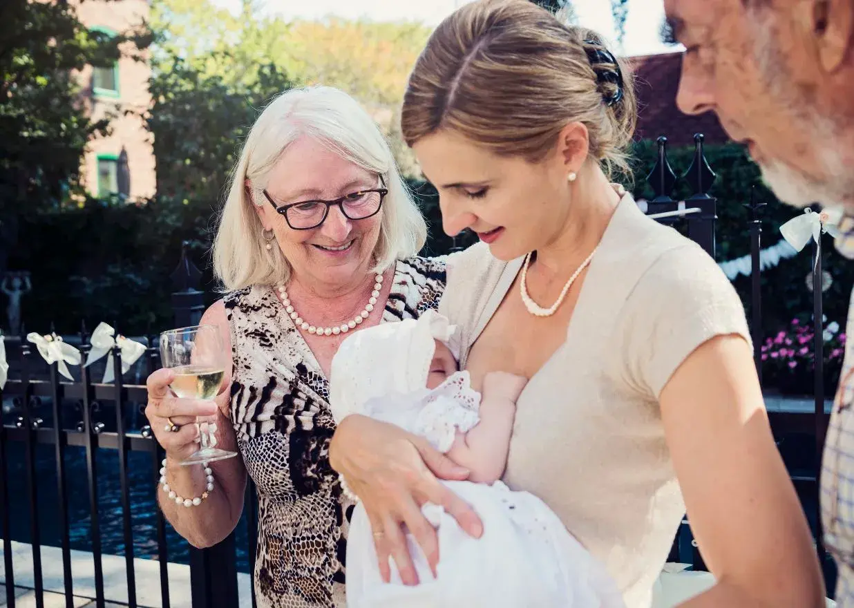 Elegancka mama karmi dziecko na chrzcinach. Babcia z kieliszkiem wina i dziadek patrzą z uśmiechem.