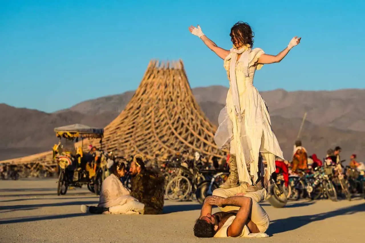 Akrobaci na pustyni podczas Burning Man. W tle charakterystyczna konstrukcja i tłum ludzi.