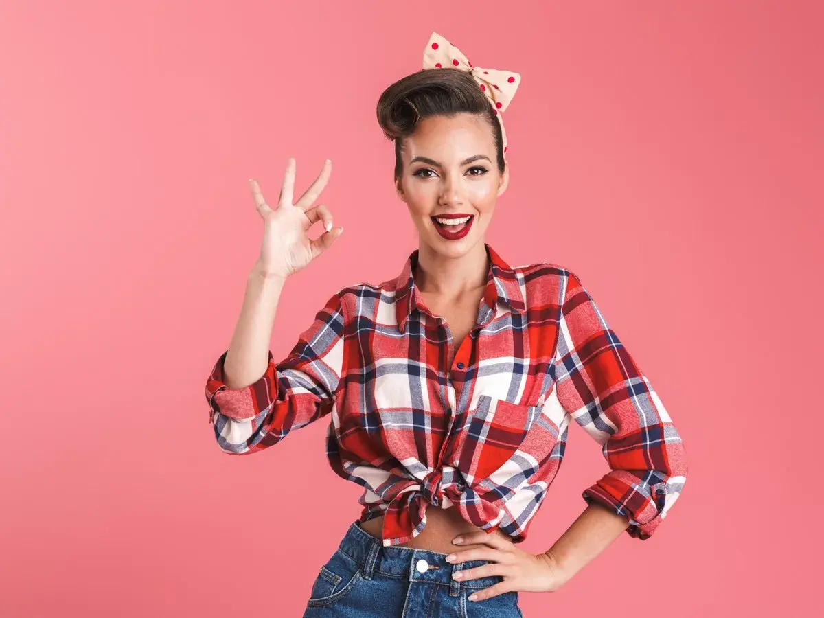 Uśmiechnięta kobieta w stylu pin up girl stylizacje, w kratę koszuli i dżinsach, pokazuje znak OK.