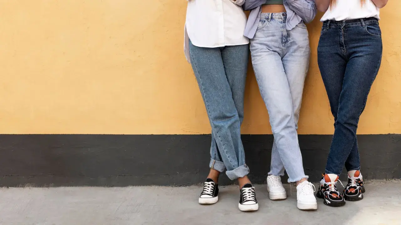 Trzy osoby w jeansach, w tym jeansy straight, stoją przy żółtej ścianie.