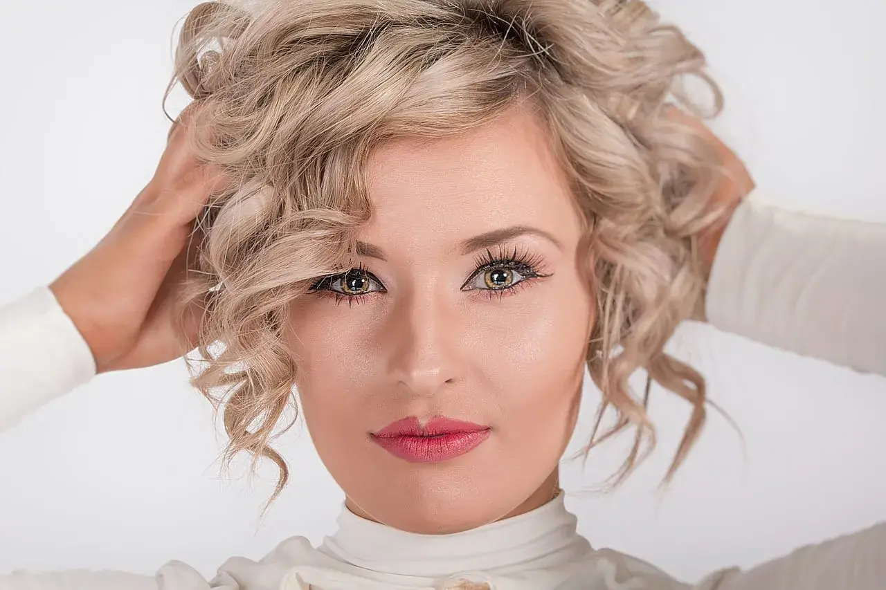Kobieta z kręconymi blond włosami, która nonszalancko układa je dłońmi. Jej spojrzenie jest hipnotyzujące, a usta pomalowane na różowo.