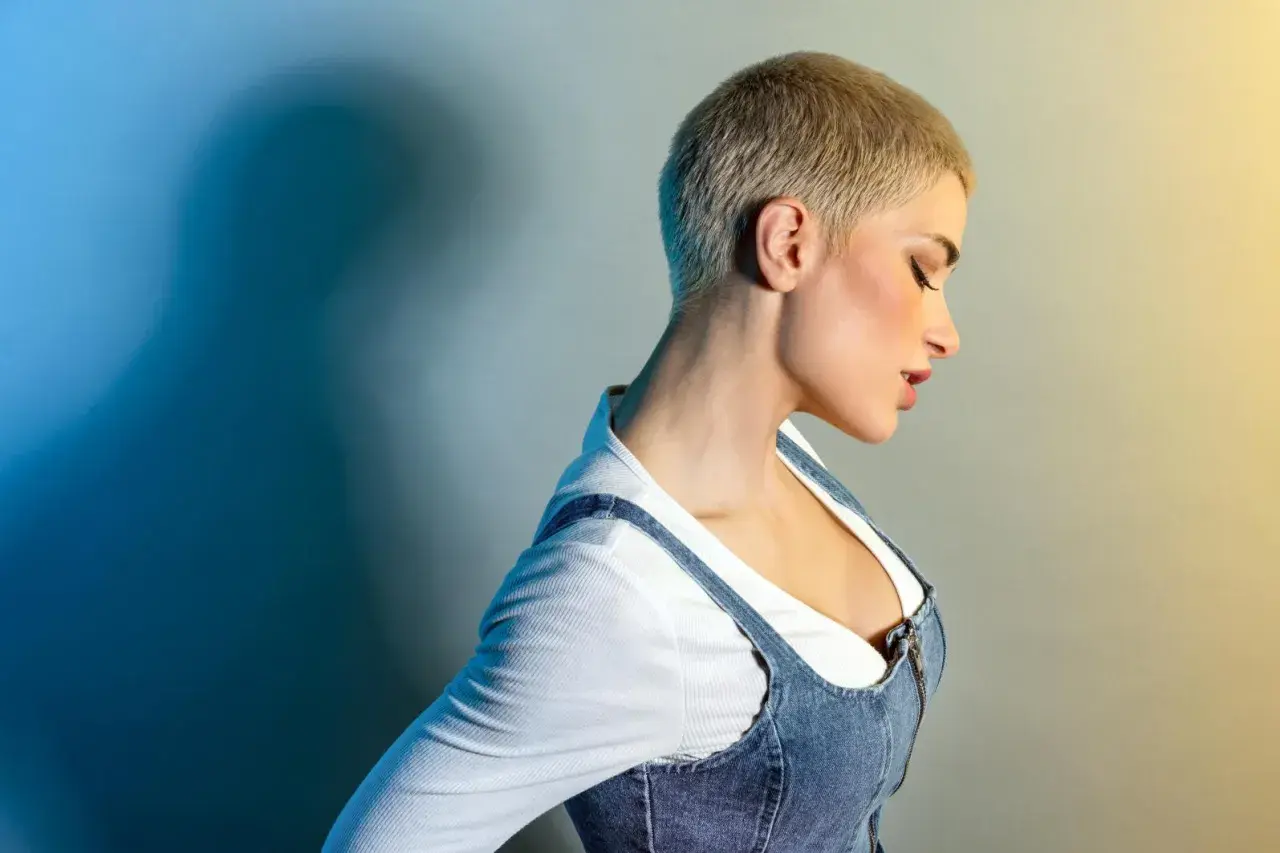 Kobieta z odważną fryzurą buzz cut, w jeansowej sukience na ramiączkach i białej bluzce.