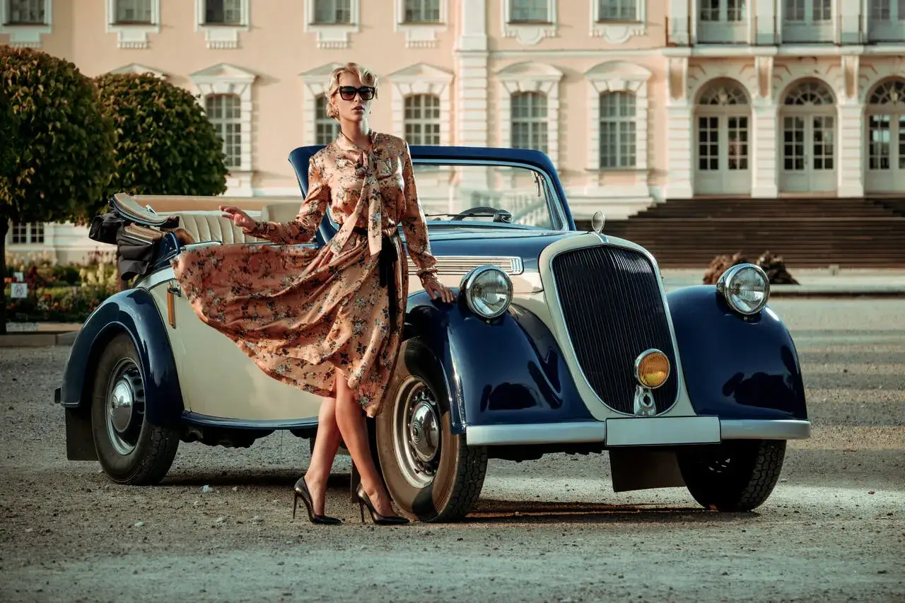 Elegancka kobieta w kwiecistej sukience i szpilkach pozuje przy zabytkowym kabriolecie. Stylowe lata 20. w pełnej krasie.