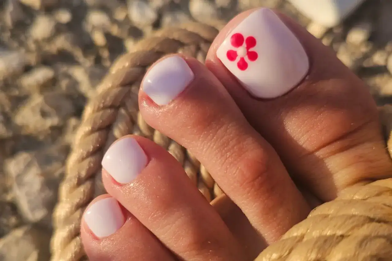 Letnie paznokcie u stóp w białym kolorze z różowym kwiatkiem. Idealne na plażę i wakacje.